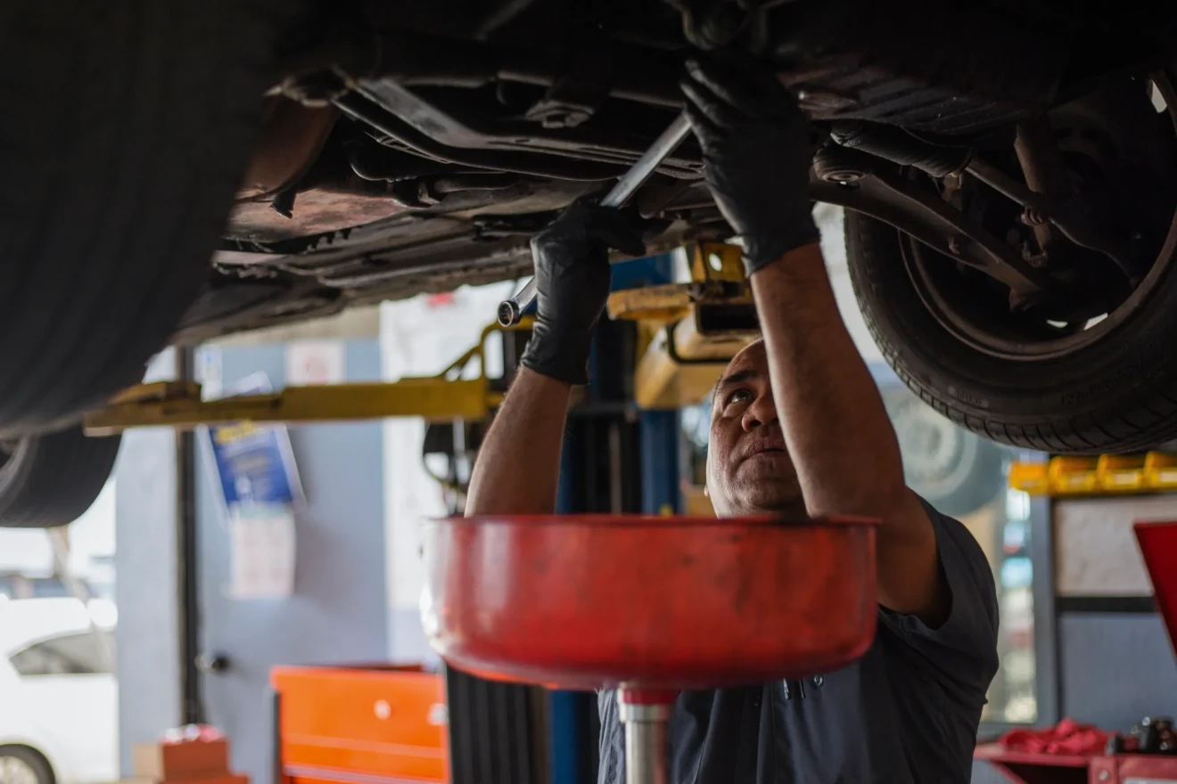 A mechanic wearing gloves looks up as he uses a tool on the underside of a raised car.